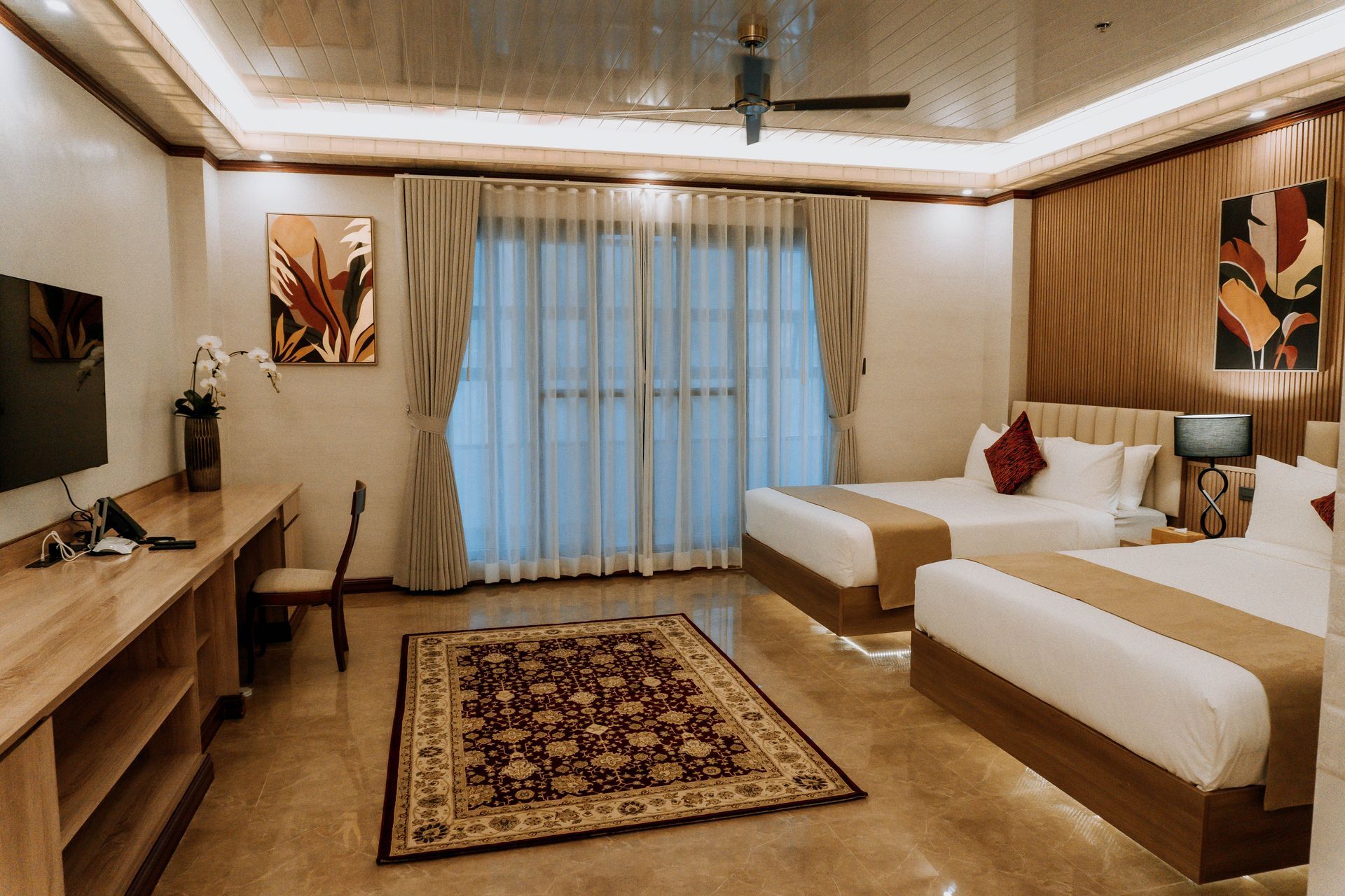 Hotel room with two beds, desk, rug, and artwork. Warm colors, wood accents, and large windows.