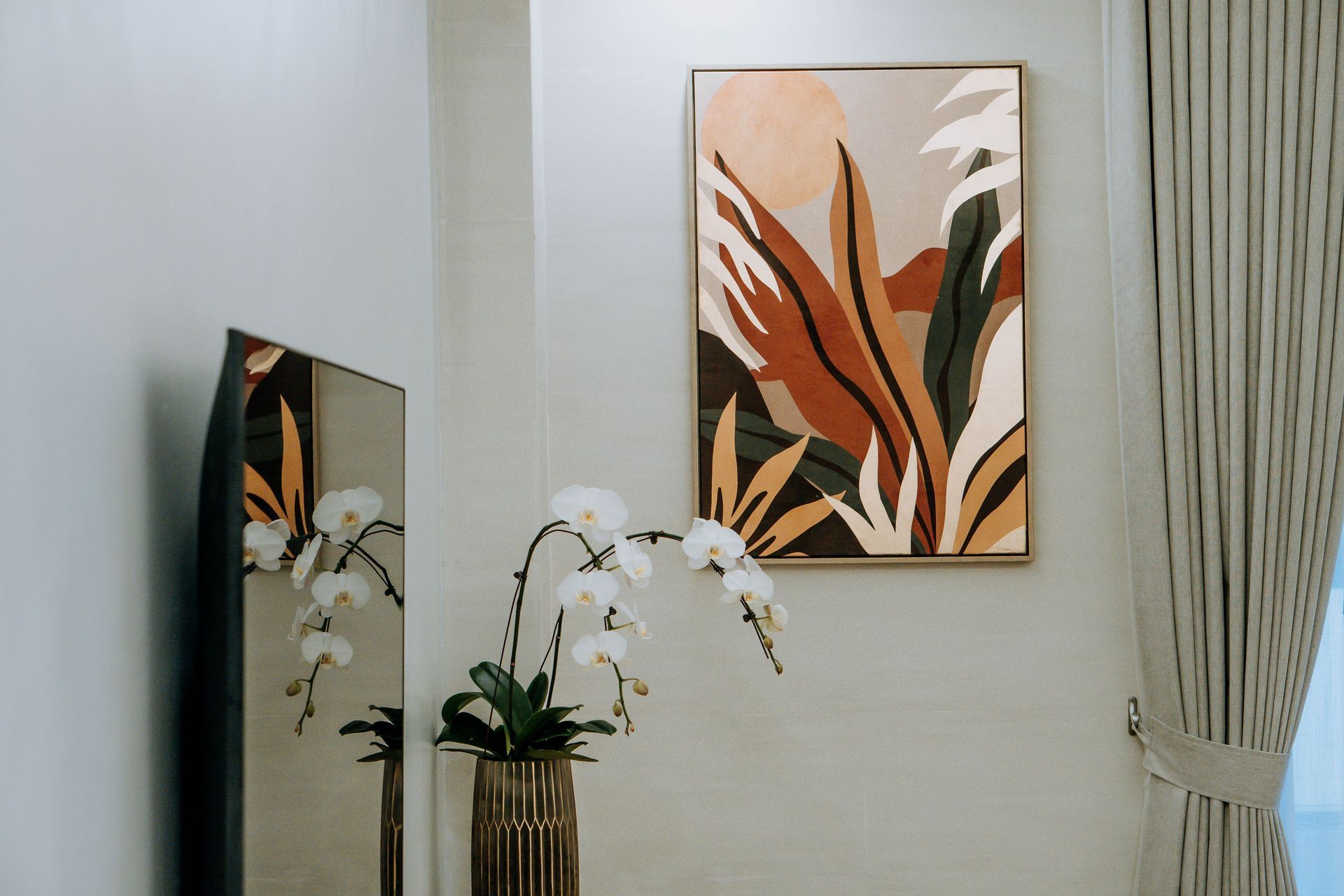 Abstract botanical art over a vase of white orchids and a tall mirror, against a neutral-toned wall.