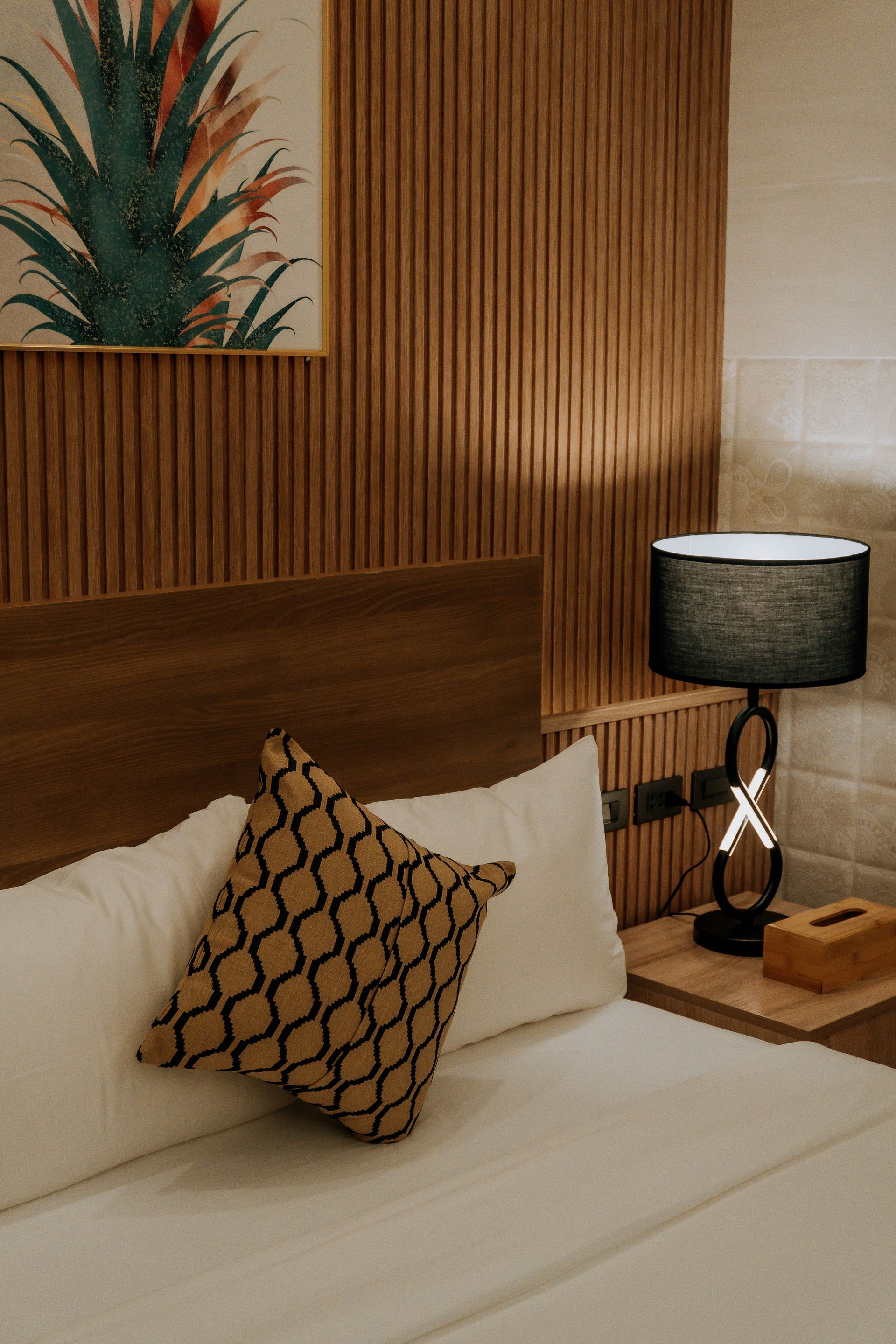 Bedroom with a wooden headboard, patterned pillow, lamp, and artwork with a plant.