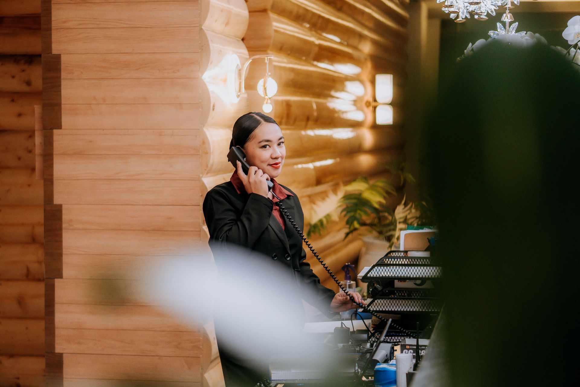 Woman in black jacket talking on a phone, standing in front of a wooden wall.