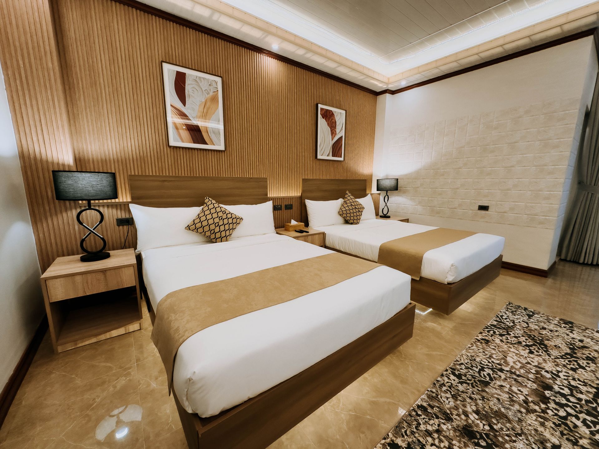 A hotel room with two beds, nightstands, lamps, and artwork, featuring wooden accents and a patterned rug.