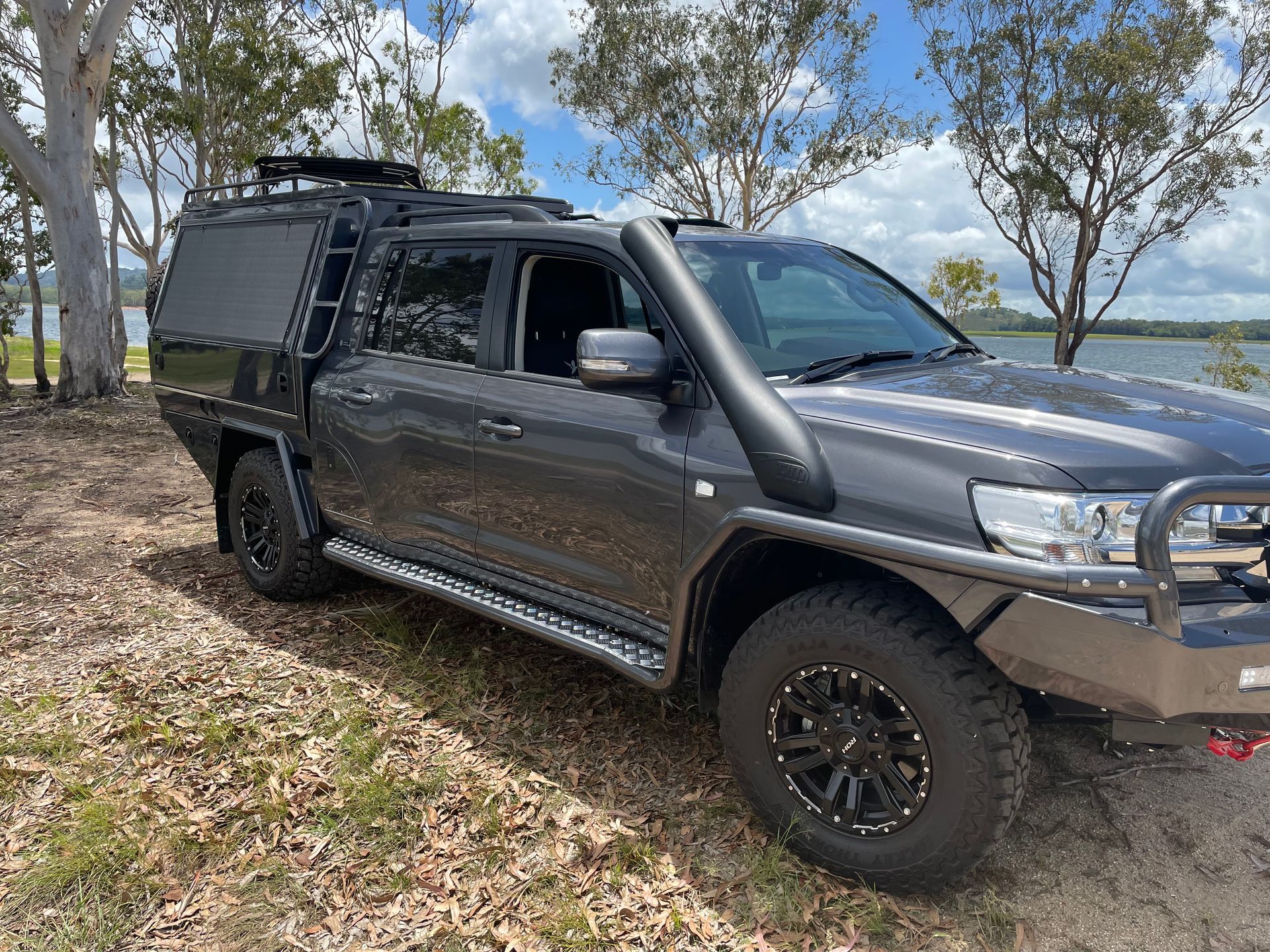 Dark gray 4x4 truck with off-road modifications parked near water. Features a snorkel, canopy, and roof rack.— Boyd's Fabrications In Portsmith, QLD 
