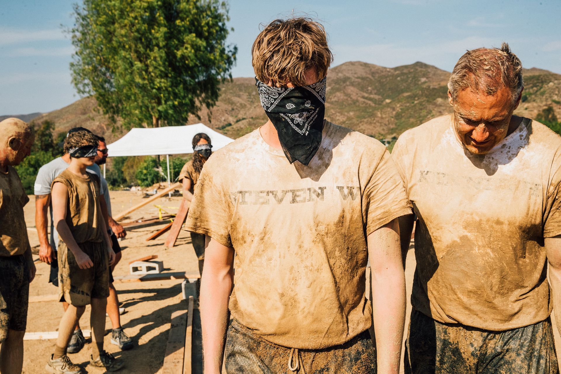 A man wearing a bandana is standing next to another man in a muddy field.