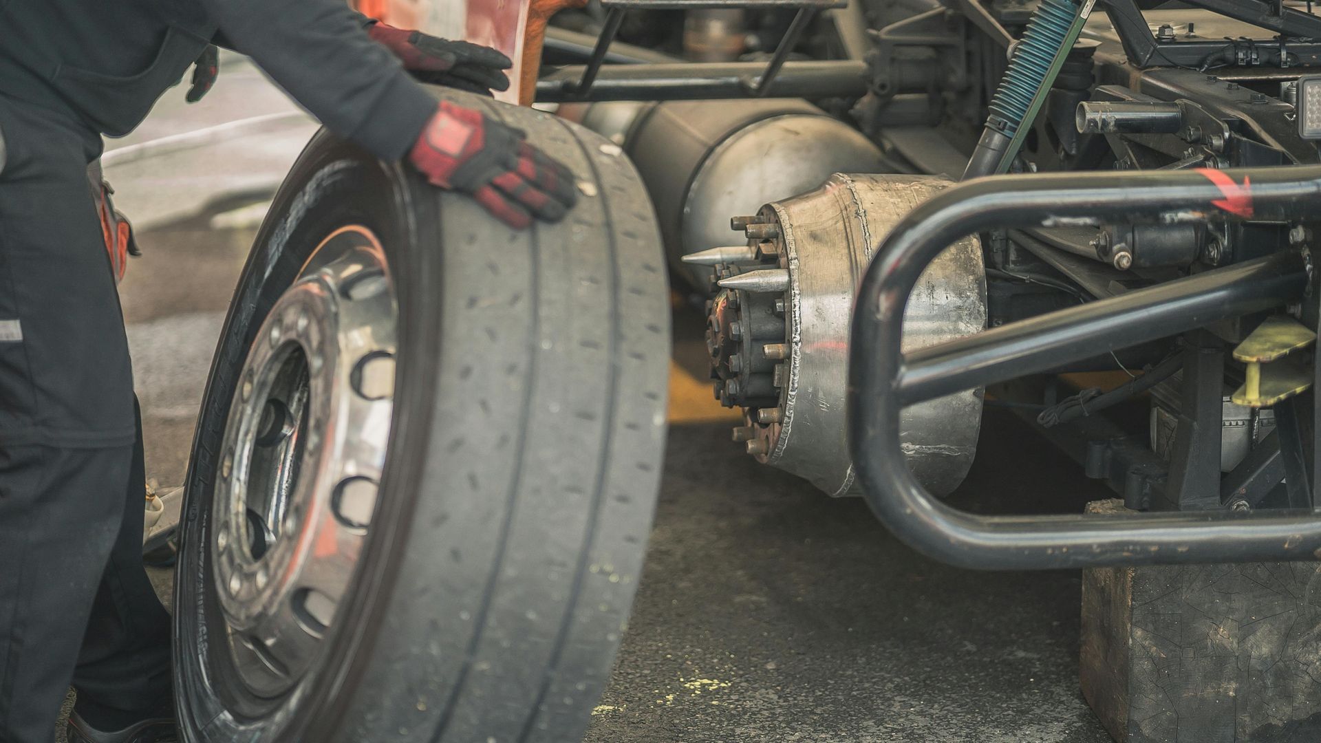 Racing car mechanic adjusting a large tire beside a race car in a garage, with gloved hands and exposed chassis.