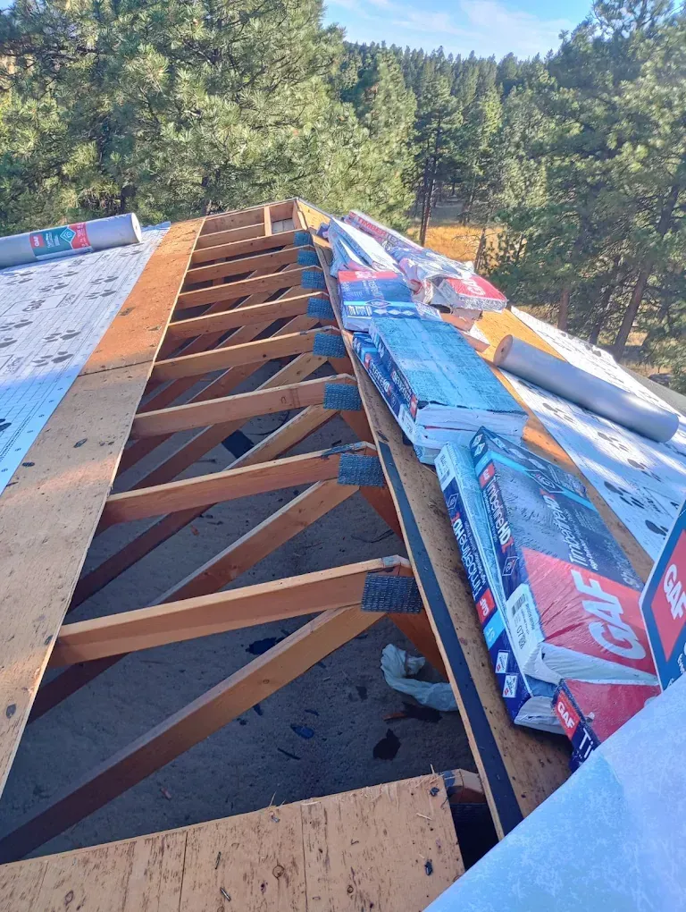 Roofing work in progress: wood trusses, stacked shingles, blue underlayment. Forest in the background.