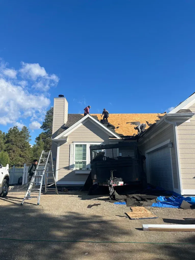 Roofers on a house removing old shingles. Blue sky, beige siding, and a black trailer.