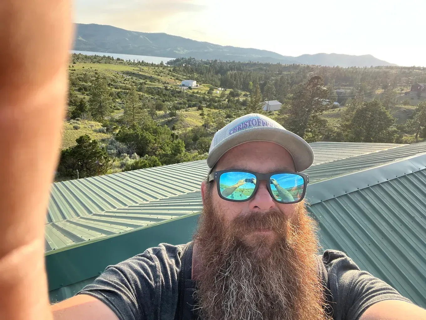 Man with a long beard and sunglasses on a rooftop, scenic mountain and lake view in the background.