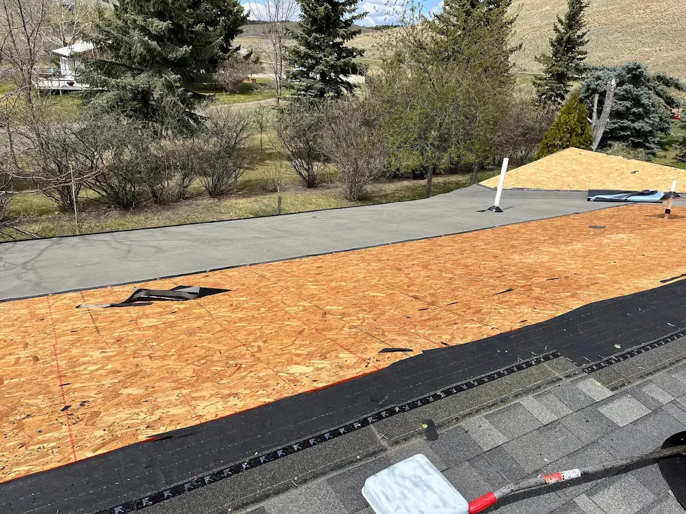 Rooftop with exposed wood panels and asphalt shingles. A driveway with trees and a pile of wood chips are in the background.