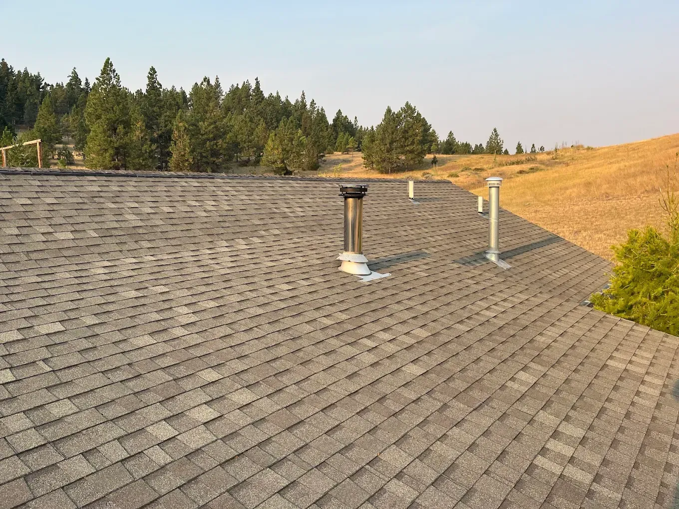 Gray shingled rooftop with multiple pipes, overlooking a field and trees.