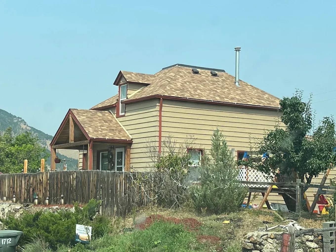 House with beige siding, brown roof, wooden fence, and a tree against a blue sky.