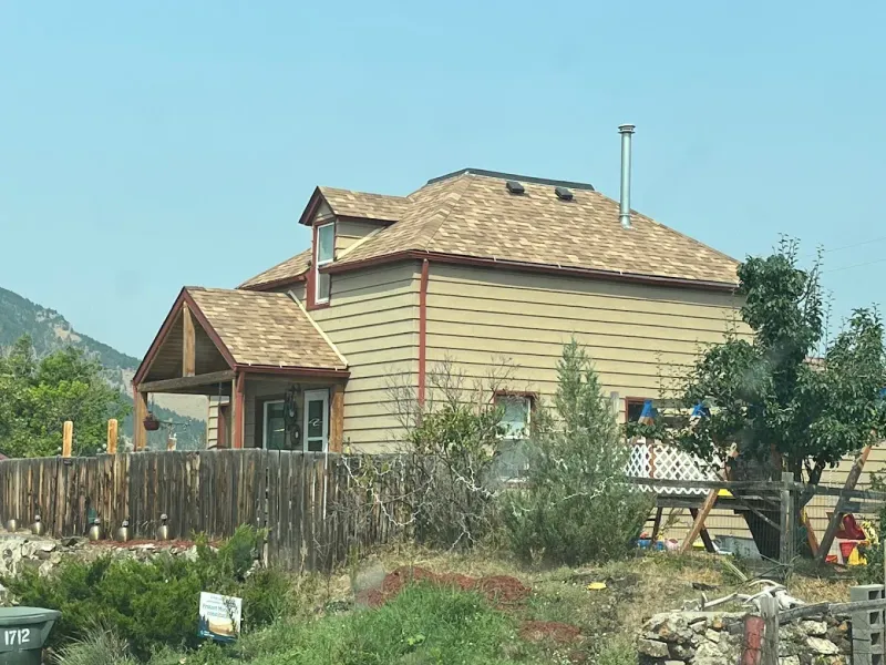 Beige house with brown roof, wooden fence, and trees against a blue sky.