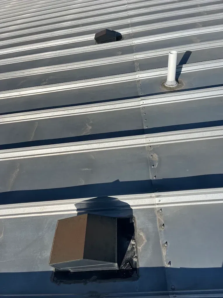 Gray metal roof with a large rectangular hole and a ventilation pipe.
