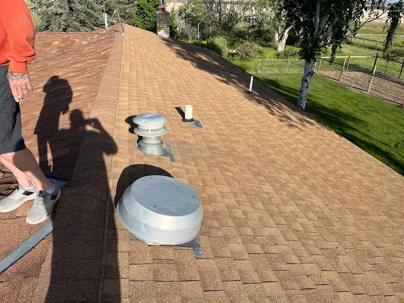Man on brown shingled roof, shadows cast. Vents and satellite dish visible, sunny outdoor setting.