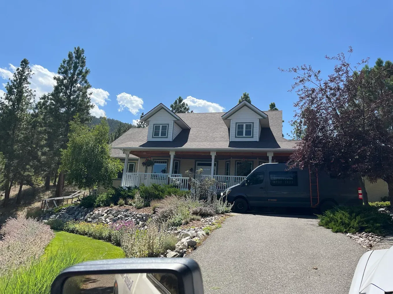 House with dormers, porch, and a gray van parked in the driveway on a sunny day.