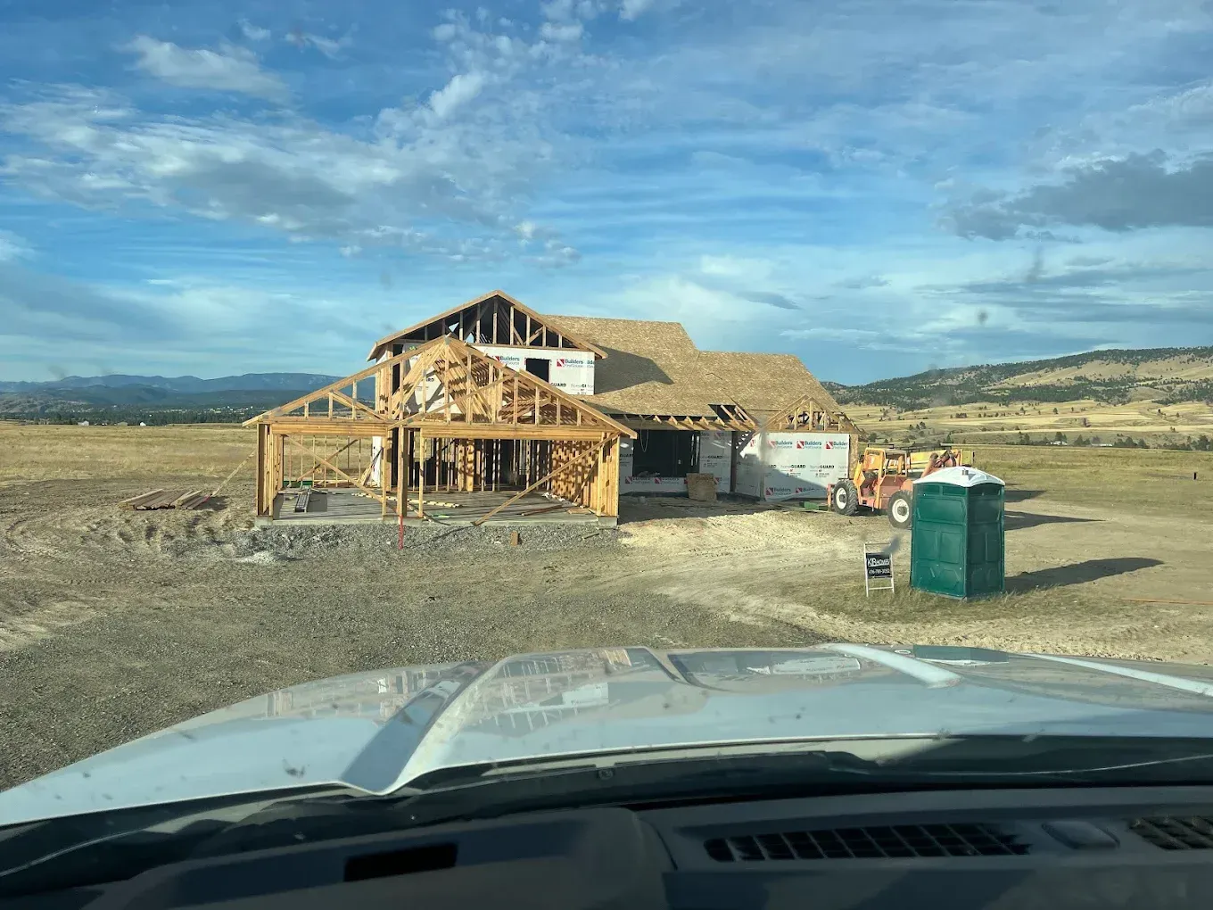 House under construction in a field, with a partly built frame, a porta-potty, and blue sky.