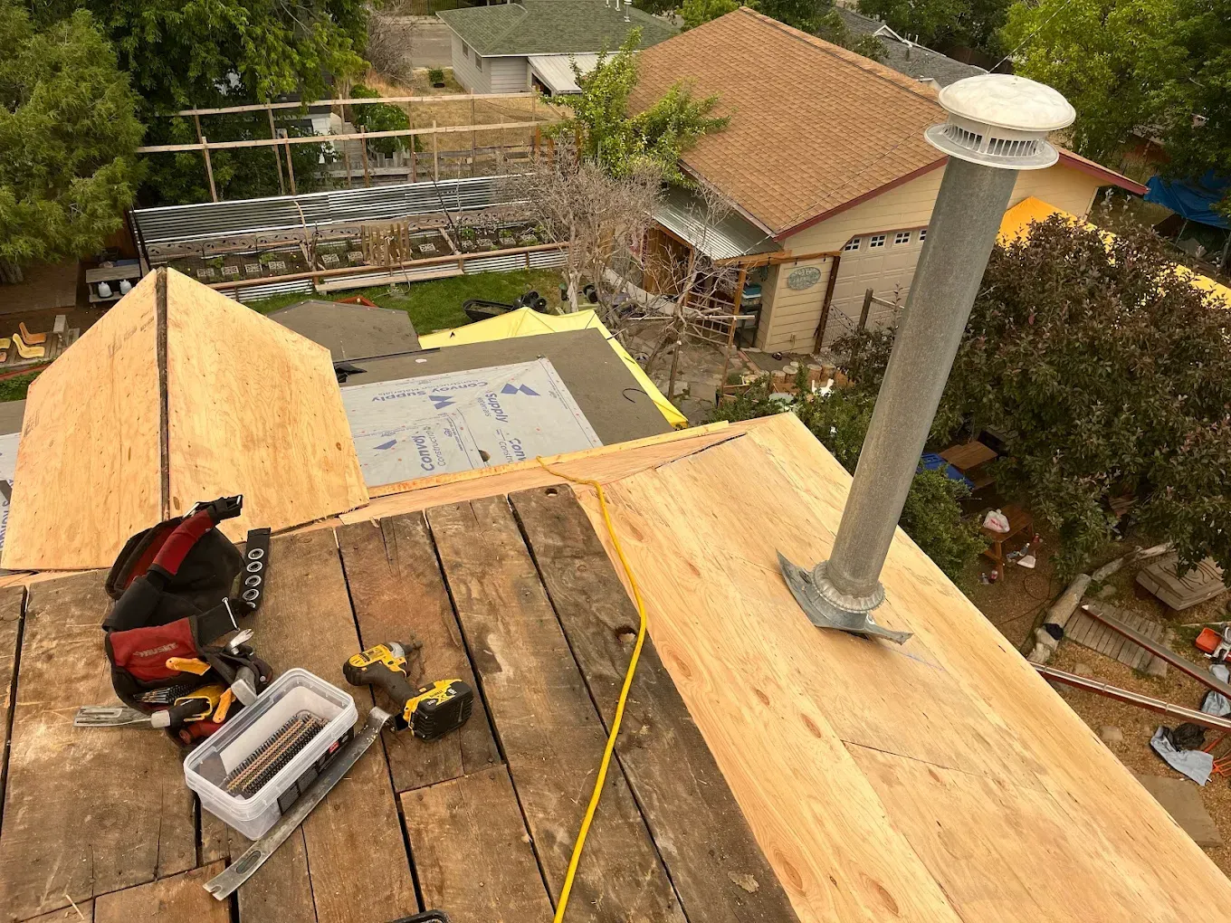 Rooftop construction scene: worker's tools, plywood, vent pipe, and surrounding houses.