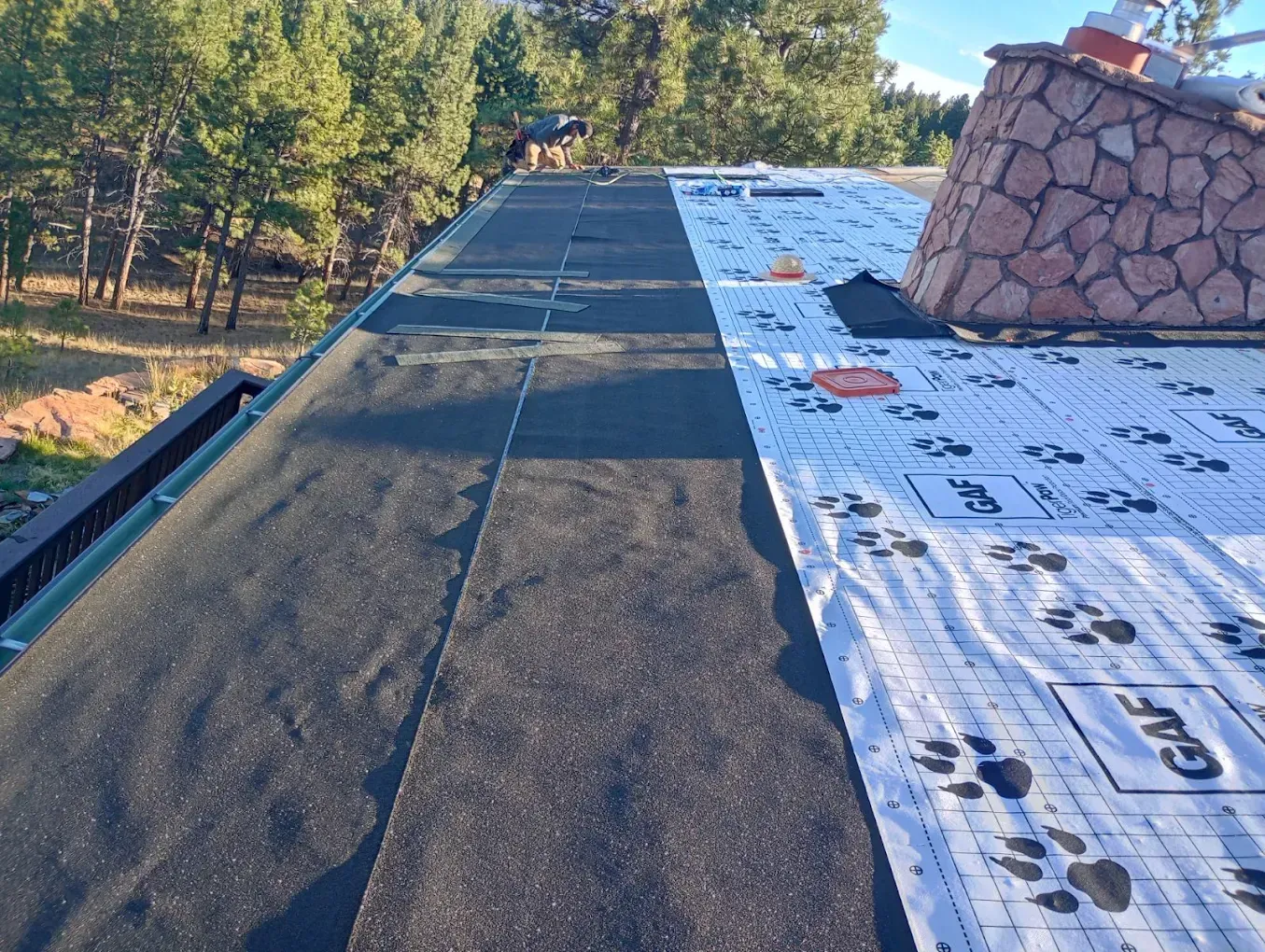 A roof partially covered with black asphalt shingles and white underlayment, near a stone chimney and trees.