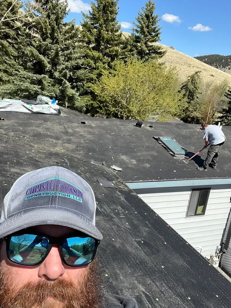 Man on roof takes selfie; another person works with a shovel, sunny day.
