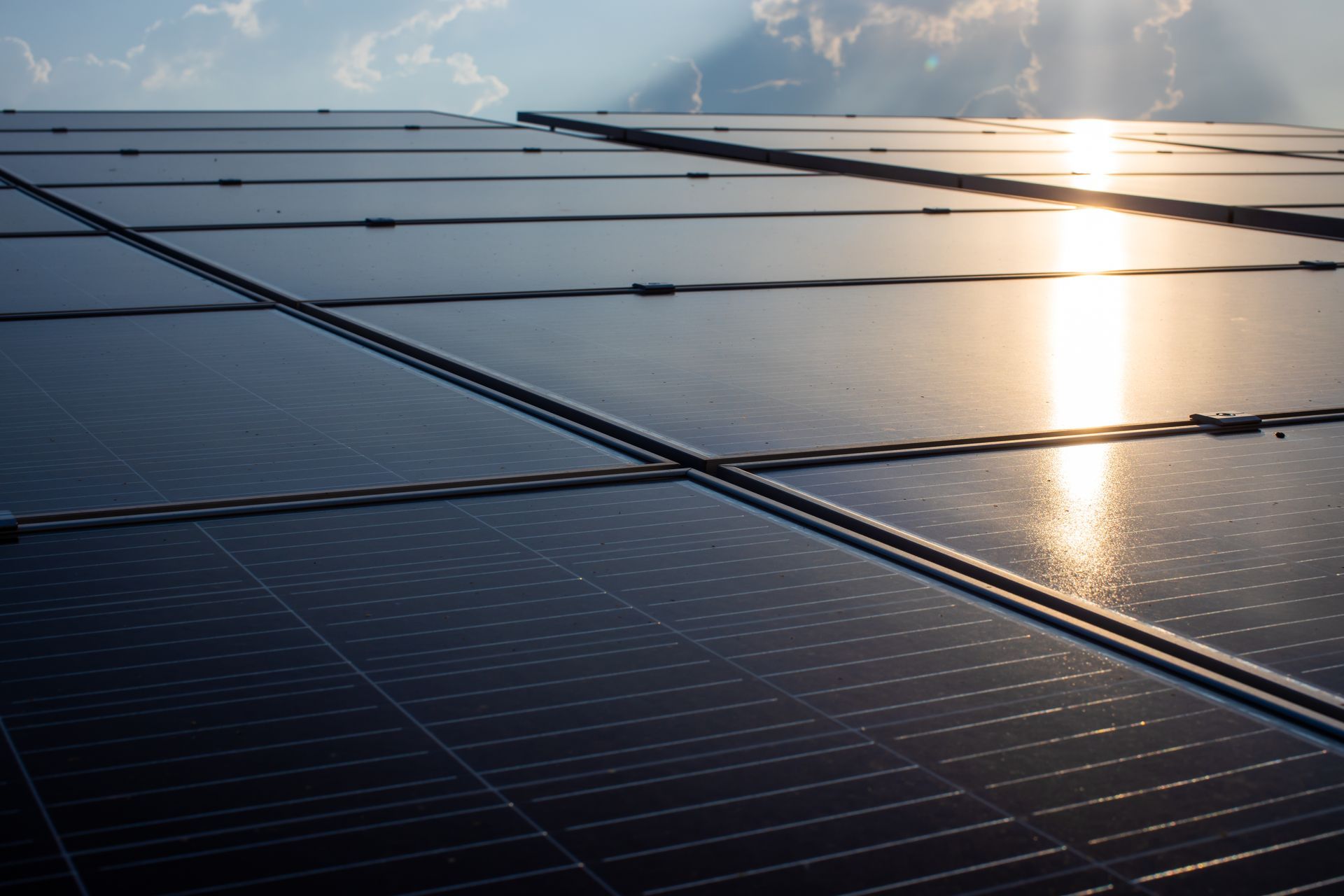 A close-up of dark solar panels reflecting bright sunlight against a sky with soft clouds.