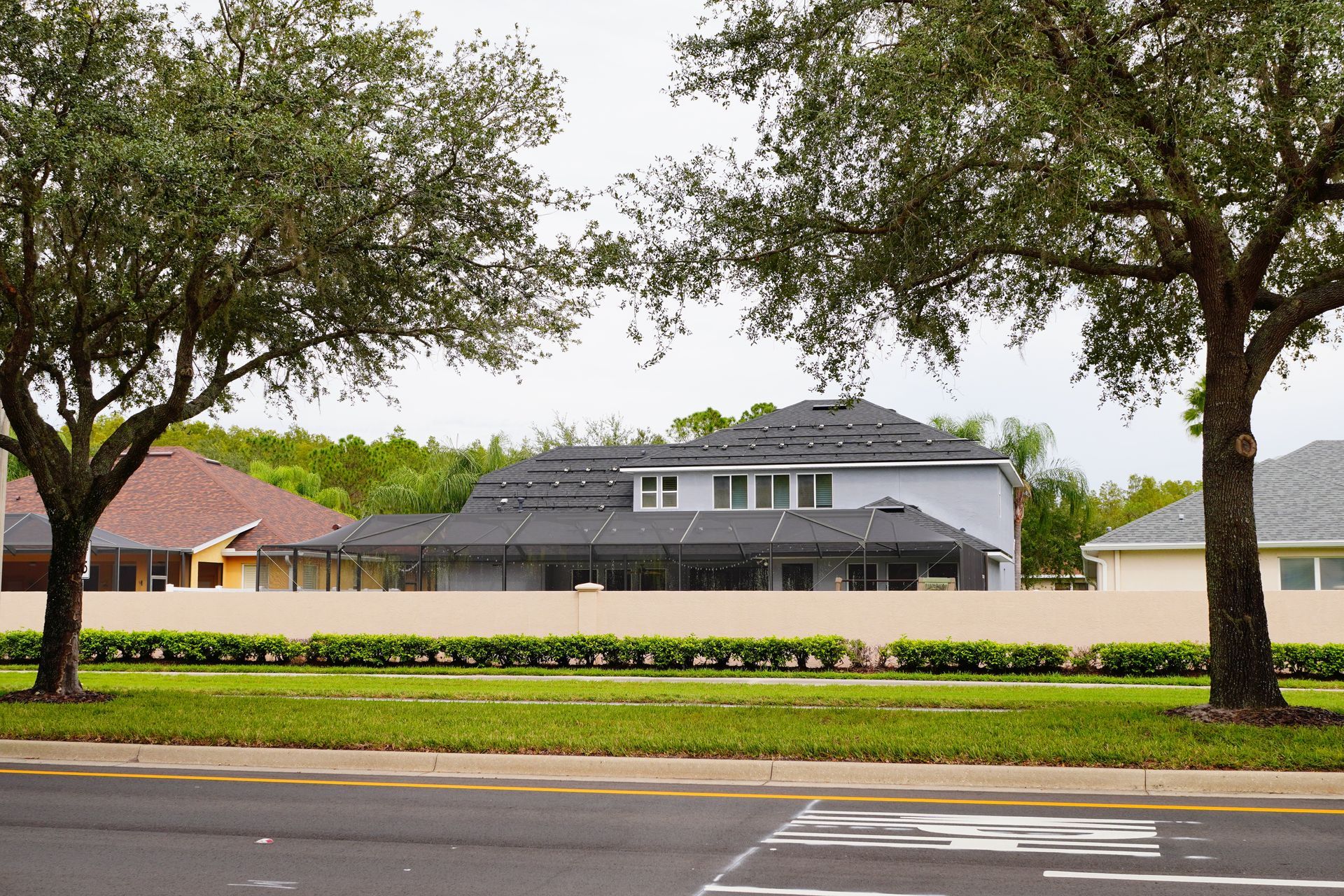 A house with a roof under construction sits behind a tan wall, framed by two large green trees and a grassy yard.