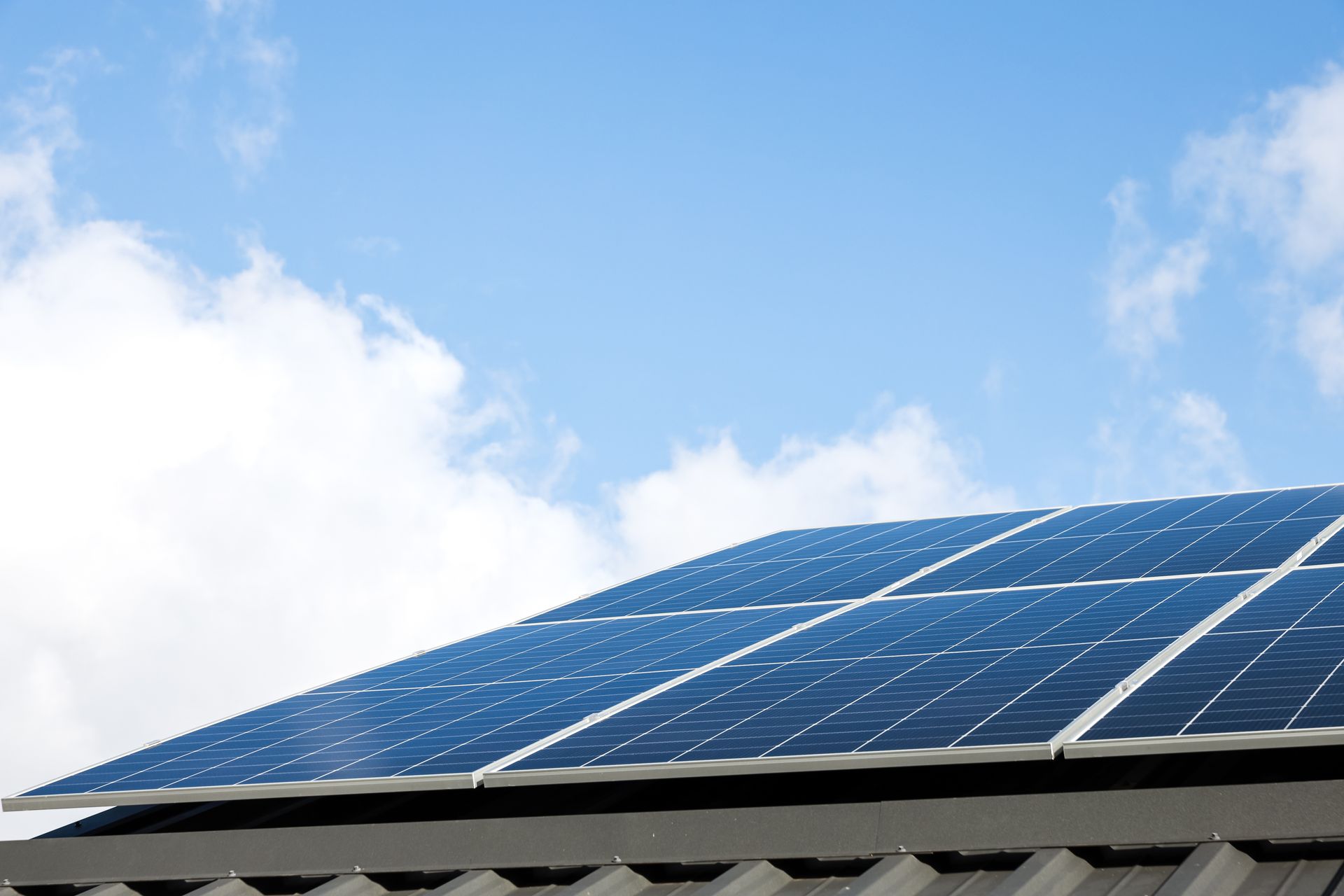 Solar panels mounted on a dark, corrugated roof against a bright blue sky with light clouds.