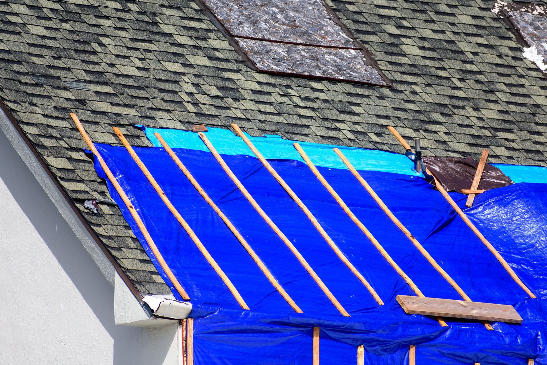 A blue tarp secured with wooden strips covers a section of a shingled roof with a skylight visible above.