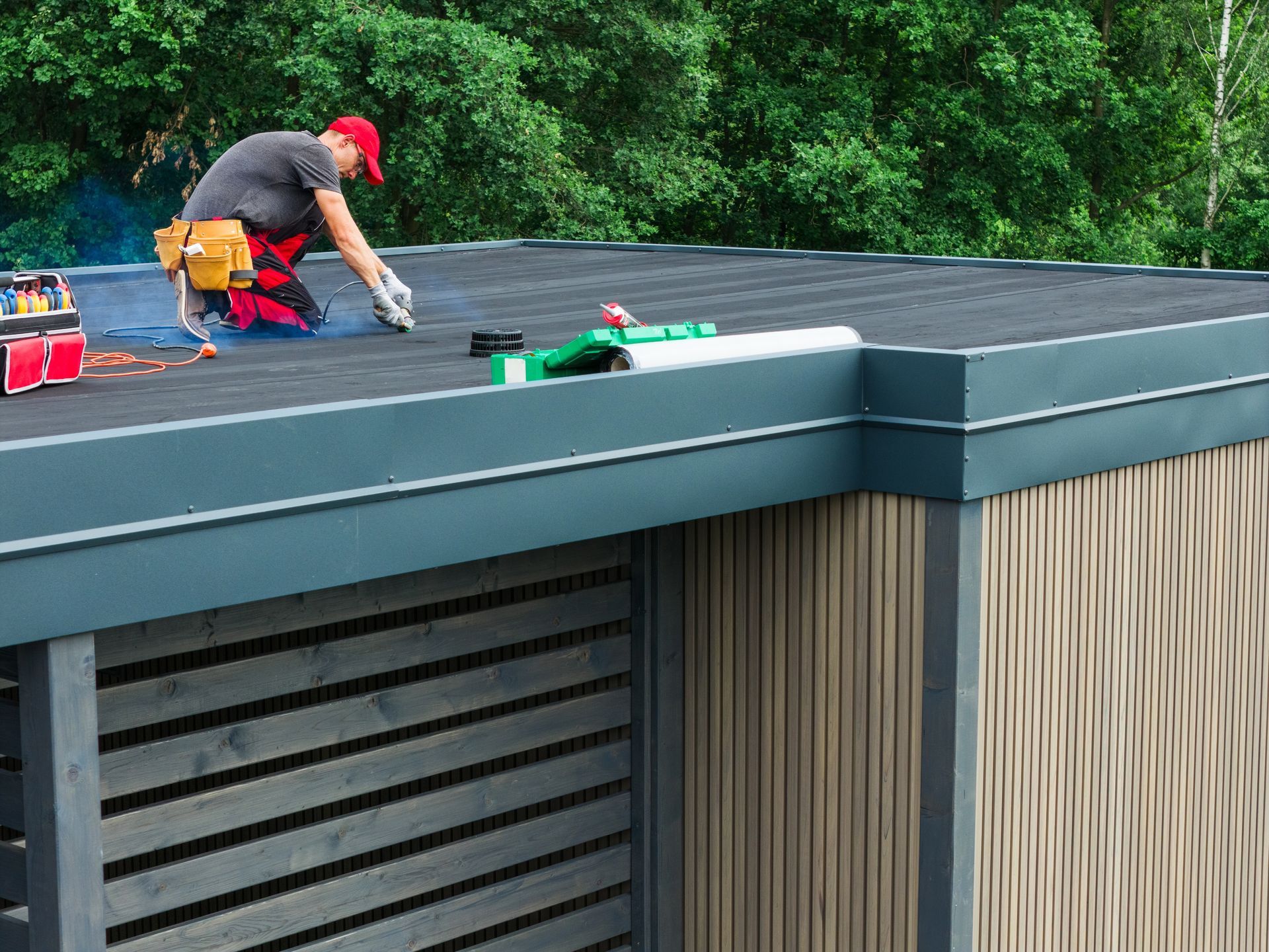 A person in a red cap and work clothes installs black roofing material on a modern flat-roofed structure with wood siding.