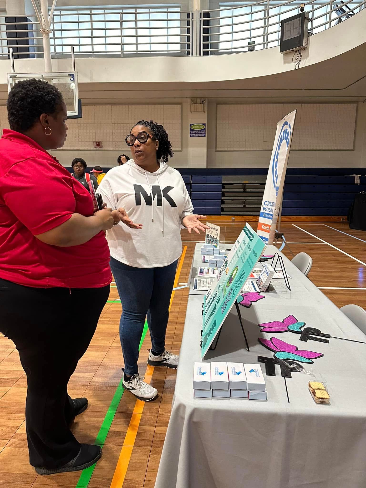 Two women are standing next to a table in a gym talking to each other.