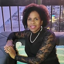 A woman in a black lace top and pearl necklace is sitting on a couch.