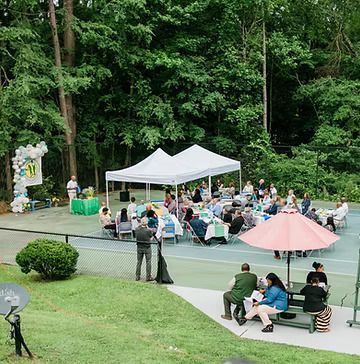 A group of people are sitting at tables under tents on a tennis court.