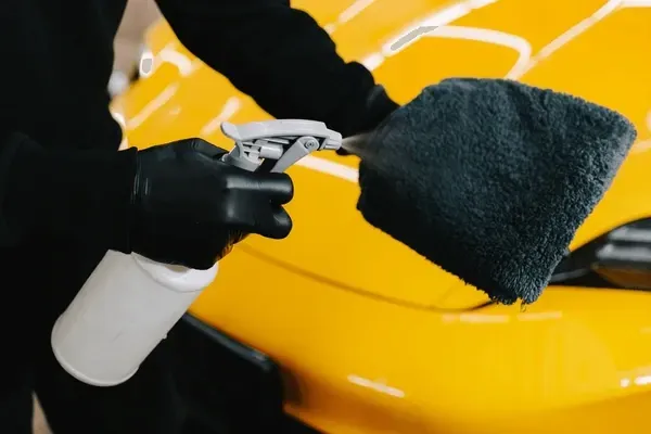Person wearing black gloves sprays cleaning product on yellow car, wiping it with a black microfiber cloth.