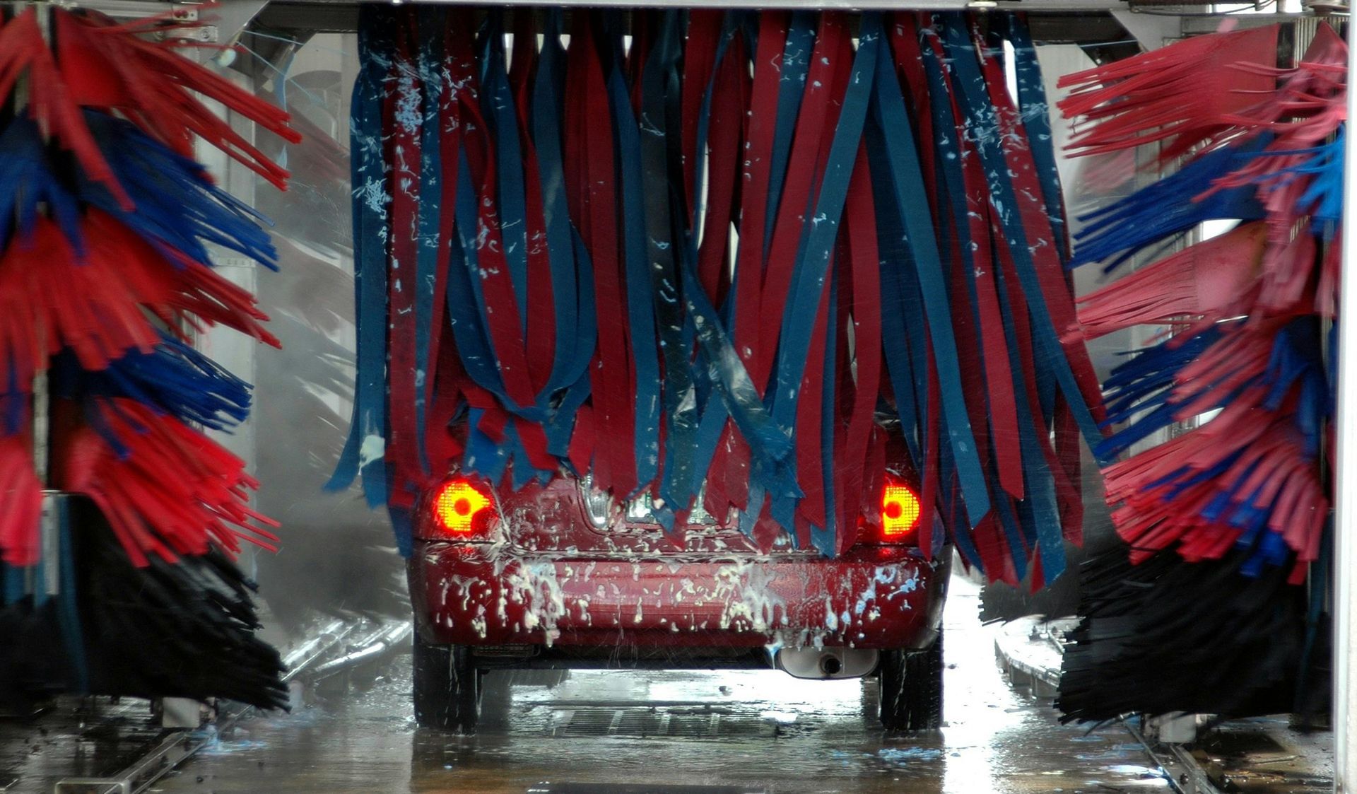 Red car in a car wash, surrounded by rotating red and blue brushes, water spraying.