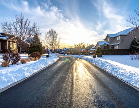 Snow-lined street in a residential neighborhood at sunrise, with wet asphalt and houses.