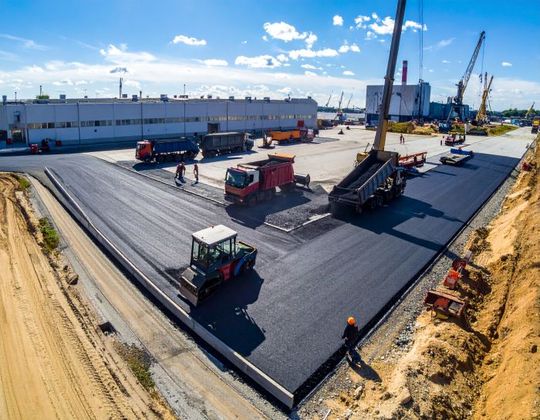 Asphalt paving in progress; trucks and workers on a construction site near a large warehouse and harbor cranes.
