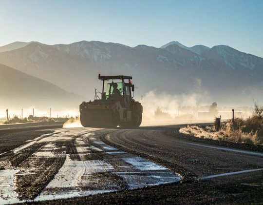Road roller compacting asphalt on a rural road with mountains in the background, a hazy sunrise.