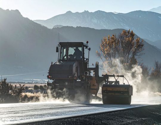 Road construction with machinery, mountains in the background, dust.