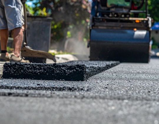 Road paving in progress, asphalt being laid and compacted by a roller. Worker with a tool in the background.