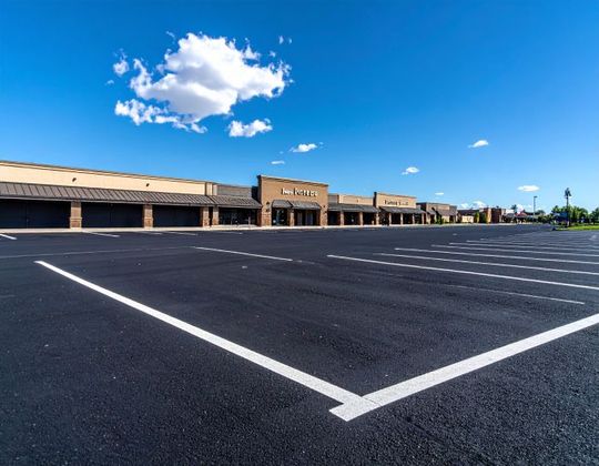 Empty asphalt parking lot in front of a strip mall under a blue sky with a single cloud.