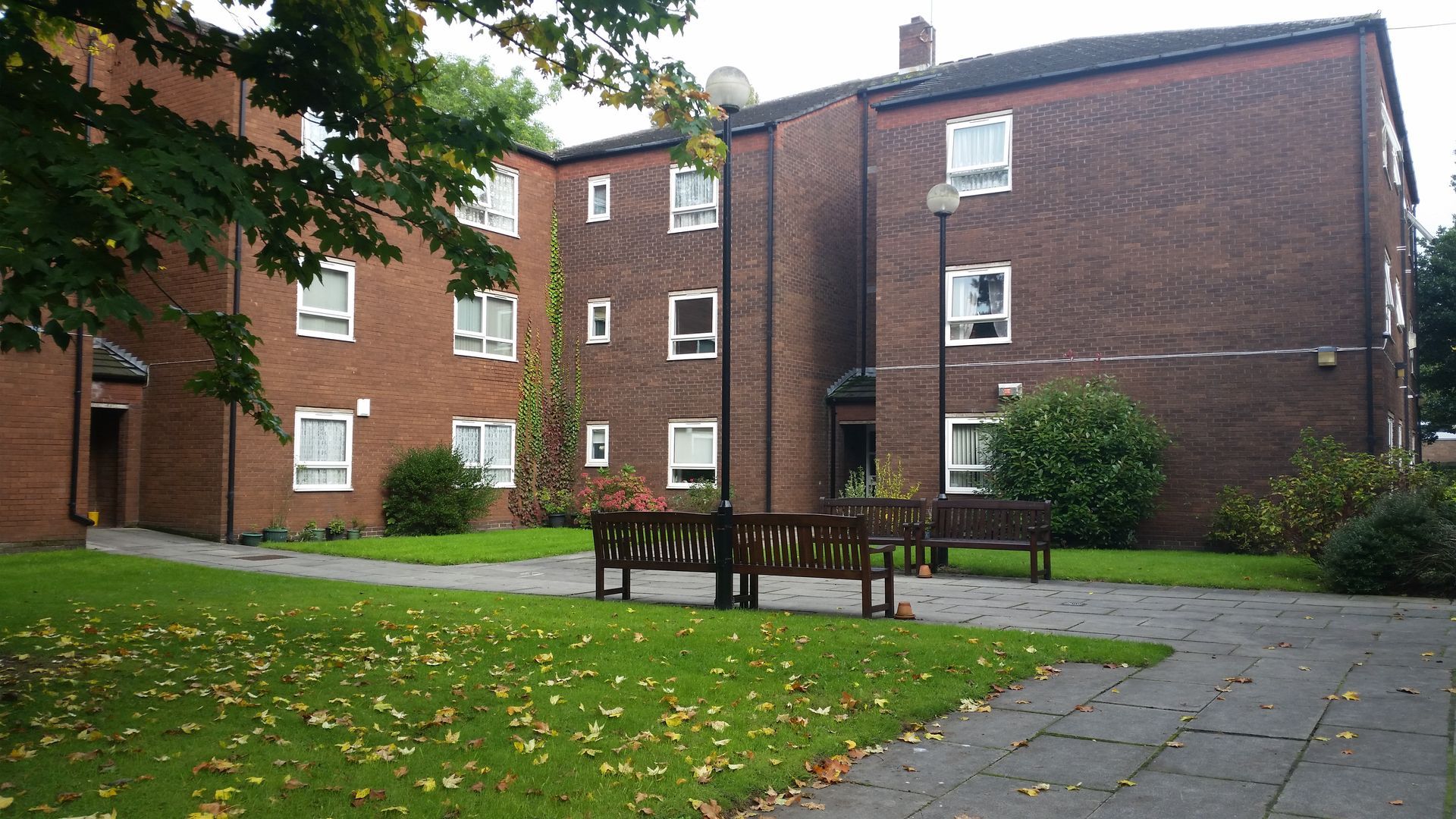 Brick apartment building with benches on a grassy area, overcast sky.