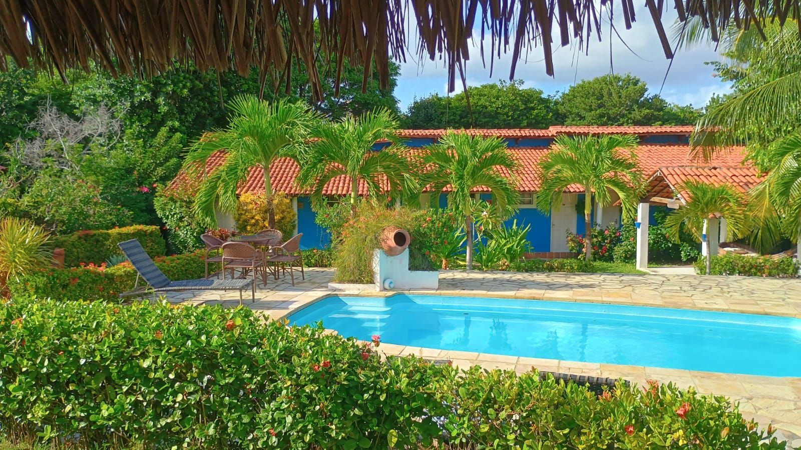 Piscina e casa tropical com persianas azuis, telhado de telha vermelha e vegetação exuberante.