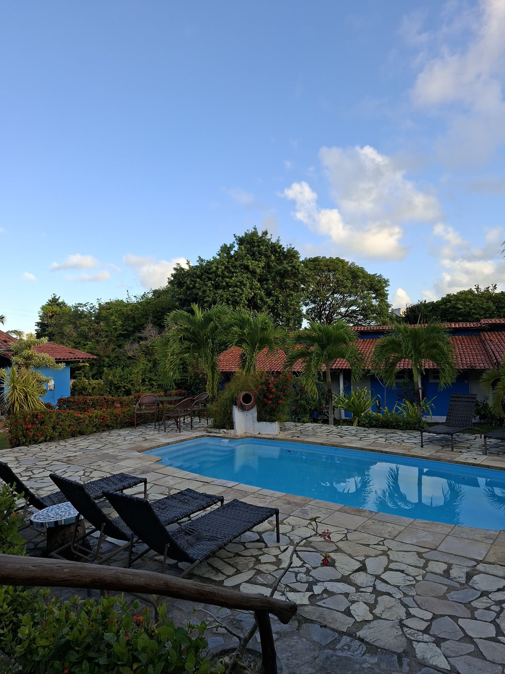 Piscina rodeada por um pátio de pedra, espreguiçadeiras, edifícios azuis e árvores verdejantes