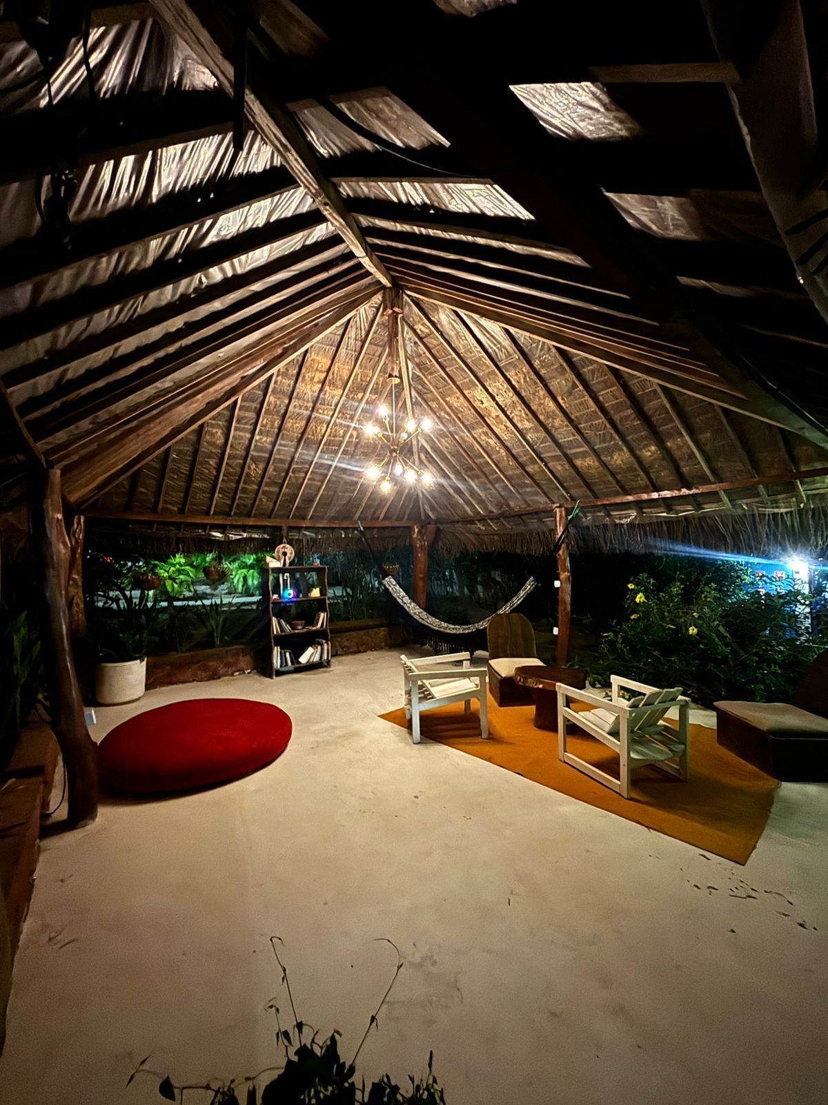 Pátio externo com teto de palha, luzes de corda, rede e assentos. Almofada vermelha no chão, tapete amarelo e folhagens.