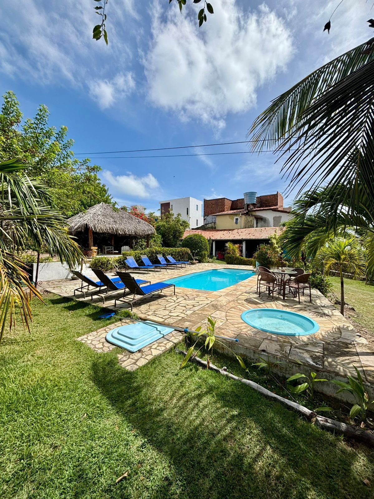 Quintal com piscina, espreguiçadeiras e um pequeno gazebo com teto de palha em um dia ensolarado.