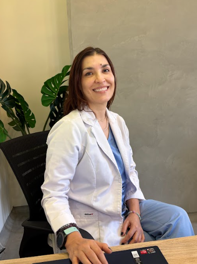 Una mujer con bata de laboratorio está sentada en un escritorio y sonriendo.