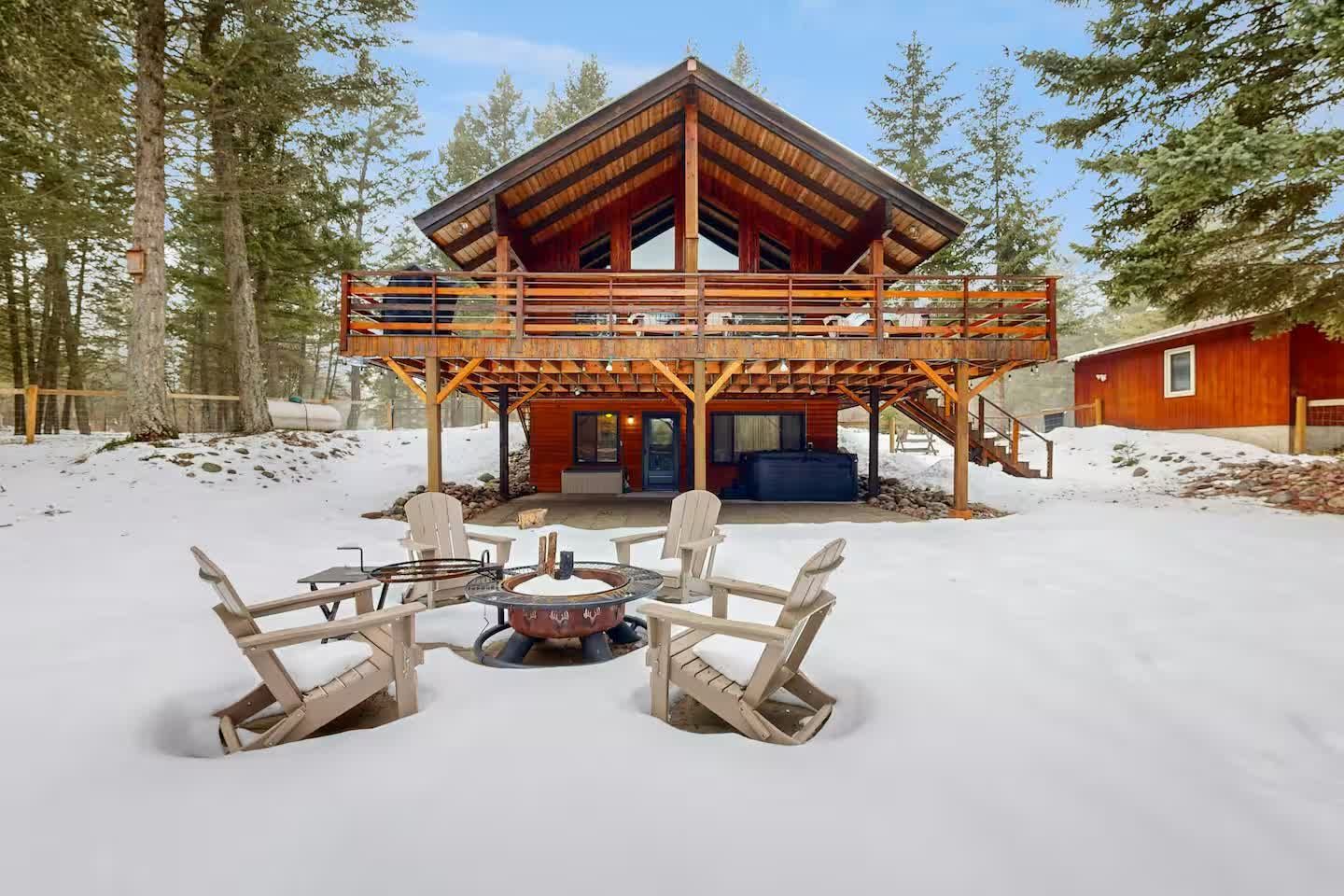 A wooden cabin with a large deck and a snowy backyard featuring a fire pit surrounded by four chairs.