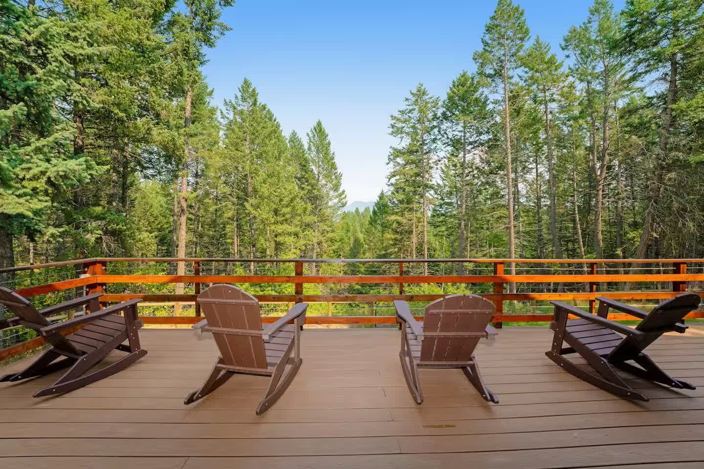 Four wooden rocking chairs sit on a deck overlooking a dense forest under a clear blue sky.