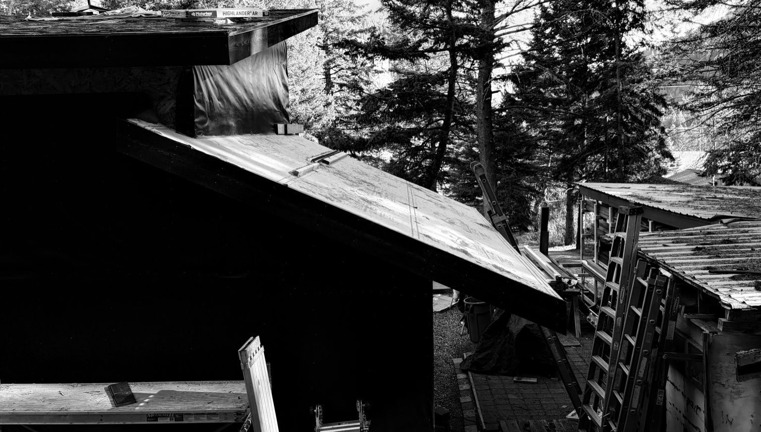 Black and white view of a dark wooden shed exterior under construction, with a ladder leaning against an adjacent structure.