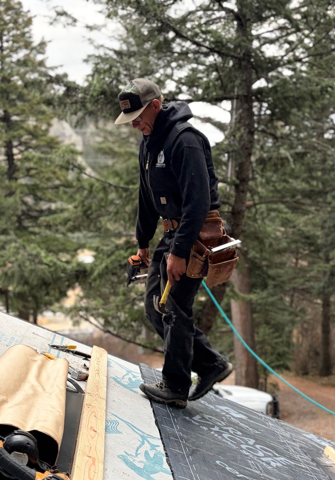 A person wearing a cap, black hoodie, and tool belt walks across a sloped residential roof near pine trees.