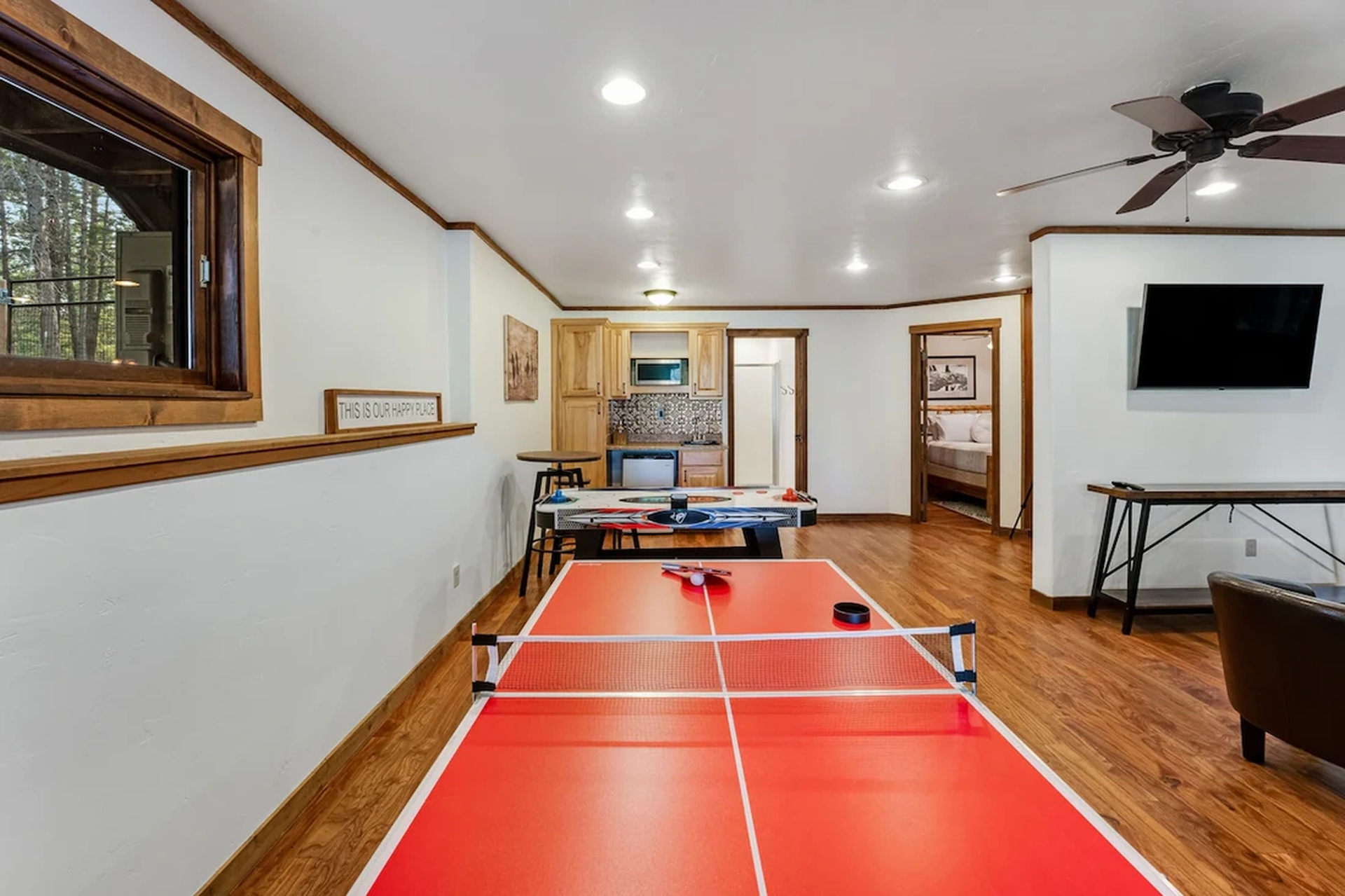 A red ping pong table in a furnished basement room with wood flooring, recessed lighting, and a wall-mounted TV.