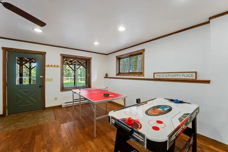 A basement game room with a red ping pong table and an air hockey table on a wood floor near two windows and a green door.